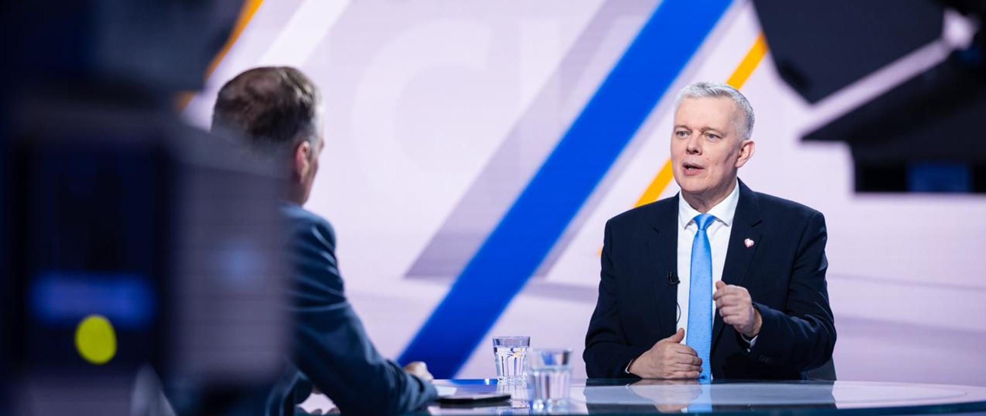 Zdjęcie przedstawia wnętrze studia telewizyjnego. Przy stole siedzą minister Tomasz Siemoniak (po prawej) i redaktor Konrad Piasecki (po lewej, tyłem do obiektywu). Na stole dwie szklanki z wodą, telefon, materiały. Po bokach zdjęcia widać fragmenty kamer i oświetlenia studia. Za ministrem szare tło z niebieskimi i żółtymi paskami. 