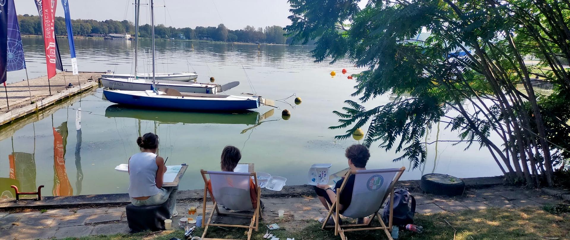 Troje uczniów siedzi na leżakach i wykonuje prawdopodobnie szkice. Jest piękna, słoneczna pogoda. Wszystko dzieje się nad Zalewem Zemborzyckim w Lublinie.