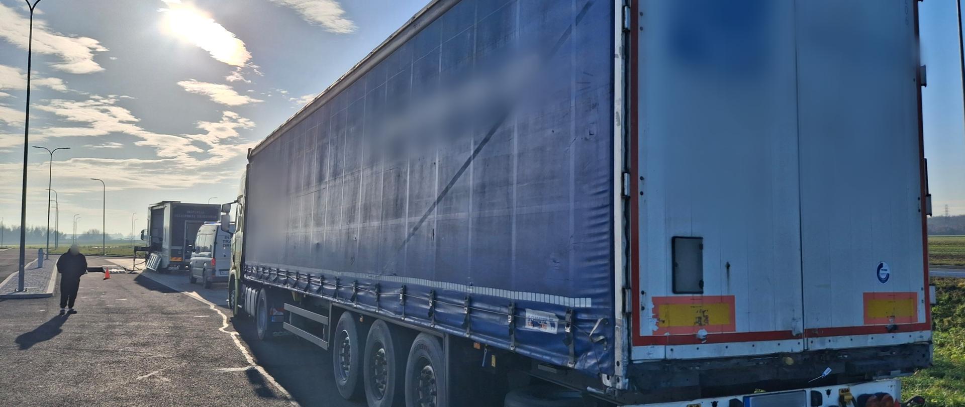 Ujęcie z tyłu i boku niebieskiej naczepy ciężarówki SCHMITZ, zaparkowanej wzdłuż parkingu. Słońce świeci intensywnie z lewej strony, tworząc jasną poświatę i długie cienie. W oddali widać kolejną ciężarówkę i idącą postać.