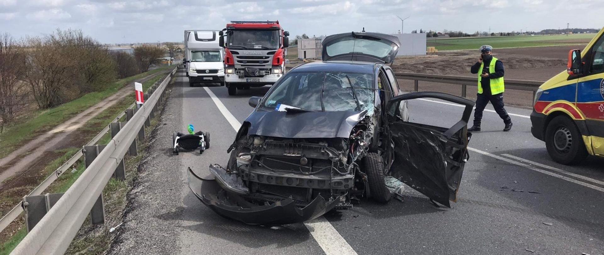 Zdjęcie przedstawiające rozbity samochód osobowy. W tle samochód pożarniczy i karetka pogotowie oraz funkcjonariusz policji wykonujący czynności służbowe