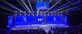 Grafika przedstawia uczestników Europejskiego Kongresu Gospodarczego oraz mówcę na scenie podczas wydarzenia. Na scenie ustawionych jest kilka ekranów wyświetlających nazwisko prowadzącego rozmowę na niebieskim tle. 