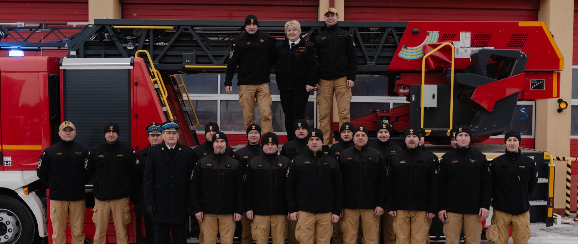 Zdjęcie przedstawia budynek Jednostki Ratowniczo-Gaśniczej Numer 3 na którego tle stoi drabina mechaniczna. Na sprzęcie stoi odchodząca funkcjonariuszka wraz z dowódcami: jednostki i zmiany na której pracowała. Poniżej pozostali strażacy, a wśród nich stoi komendant miejski PSP w Kielcach.