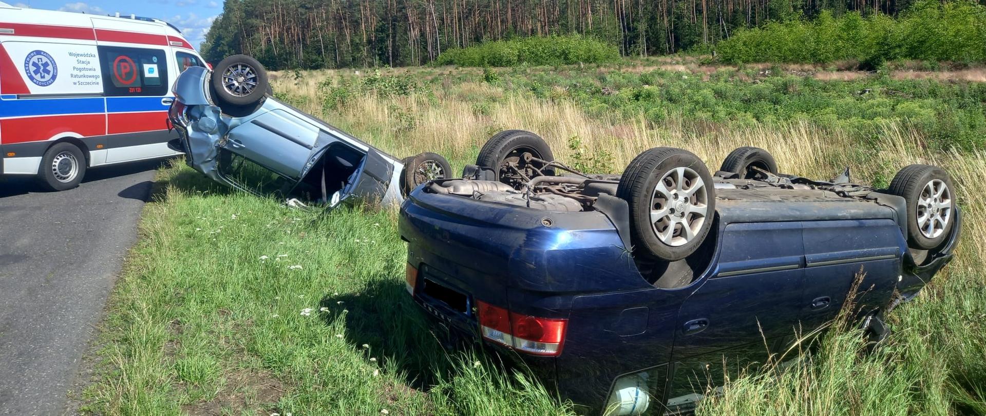 Zdjęcie przedstawia pobocze drogi na której na dachu leżą dwa samochody osobowe obok nich znajduje się karetka pogotowia w tle widzimy las.