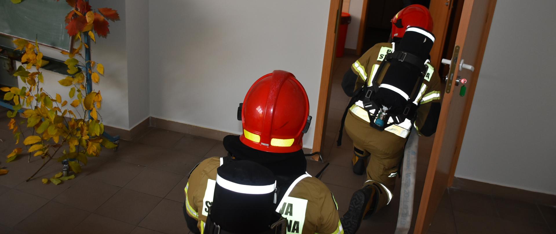 Zdjęcie przedstawia korytarz szpitala. Dwóch strażaków w pełnym umundurowaniu specjalnym oraz pracujący w aparatach ochrony dróg oddechowych udający się do strefy zagrożenia, wchodzący do pomieszczenia gospodarczego.