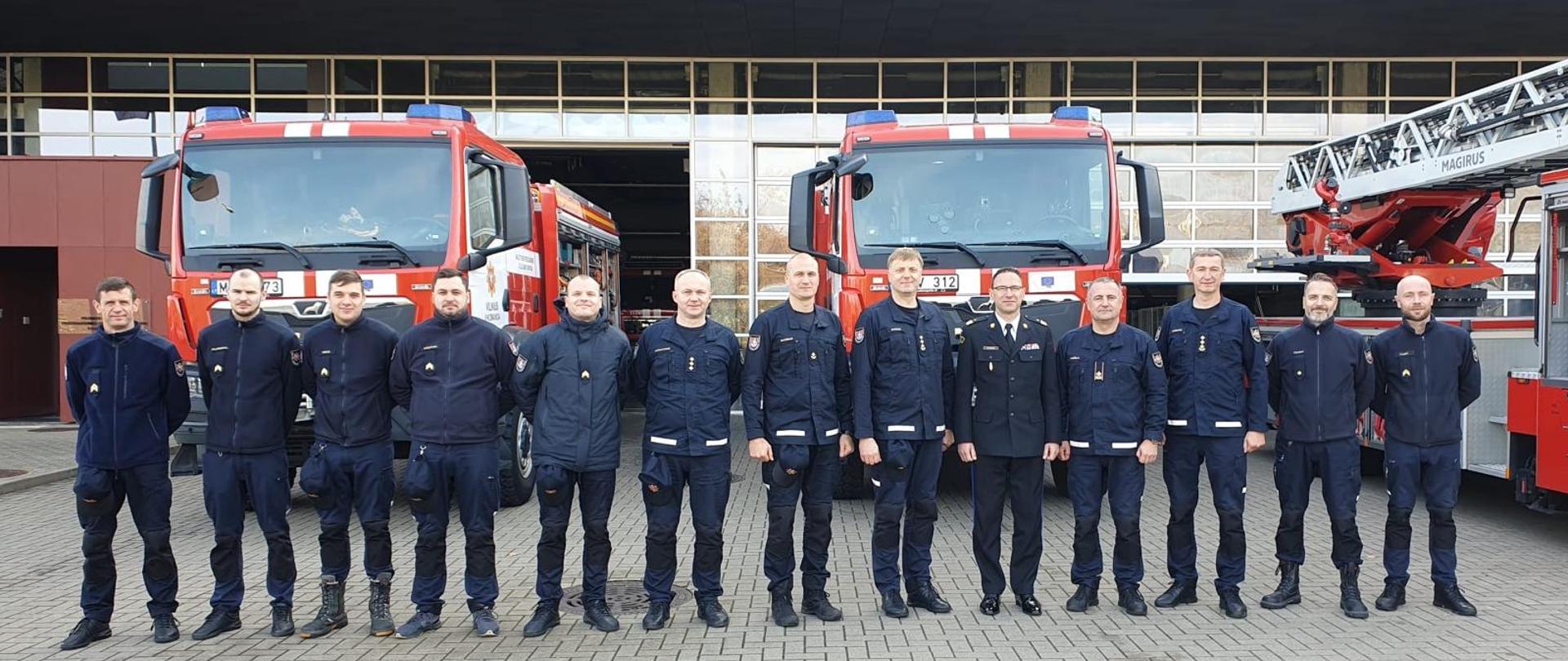 Grupa strażaków stoi w jednym szeregu przed dwoma czerwonymi samochodami ratowniczo-gaśniczymi. W tle widać budynek jednostki ratowniczo-gaśniczej z otwartymi bramami garażowymi.
