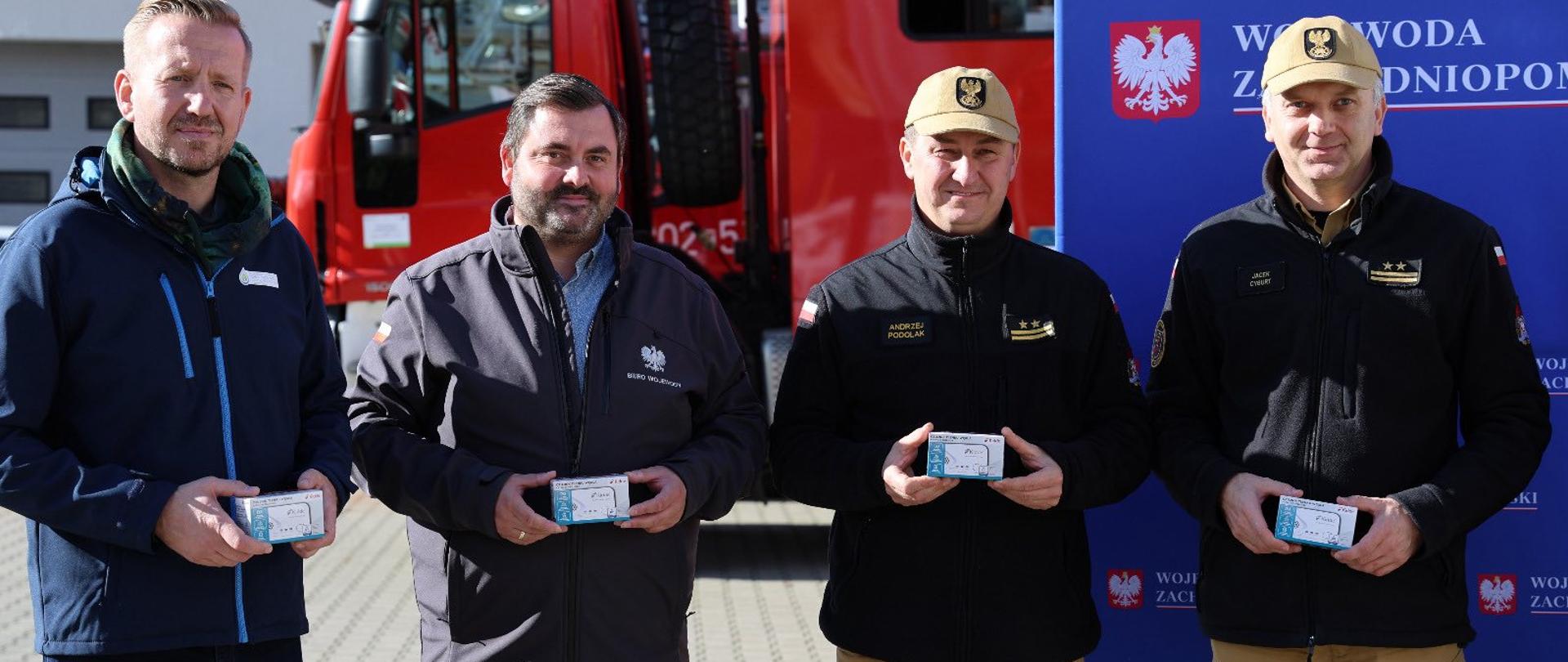 Na zdjęciu widać czterech mężczyzn stojących na zewnątrz w słoneczny dzień. Za nimi znajduje się czerwony wóz strażacki oraz niebieska ścianka z logo i napisem „Wojewoda Zachodniopomorski”. Wszyscy mężczyźni trzymają w dłoniach małe pudełka – wyglądają jak upominki lub wyróżnienia. Dwóch z nich ubranych jest w piaskowe spodnie i czarne bluzy z naszywkami, a także jasne czapki z godłem. Pozostali dwaj mają ciemne kurtki – jeden granatową, drugi szarą – obaj bez nakryć głowy. Wszyscy stoją obok siebie i patrzą w stronę obiektywu, lekko się uśmiechając.