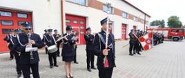 Zdjęcie zrobione na placu kętrzyńskiej komendy straży pożarnej. W szeregu ustawieni strażacy w czarnych mundurach galowych. Z lewej strony orkiestra z instrumentami, po prawej pododdział ze sztandarem. W tle czerwony samochód strażacki.