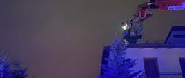 Zdjęcie w porze nocnej. Na zdjęciu budynek mieszkalny. Budynek otynkowany. Przy budynku drzewa i krzewy. Widać samochód z podnośnikiem. Pojazd straży pożarnej,