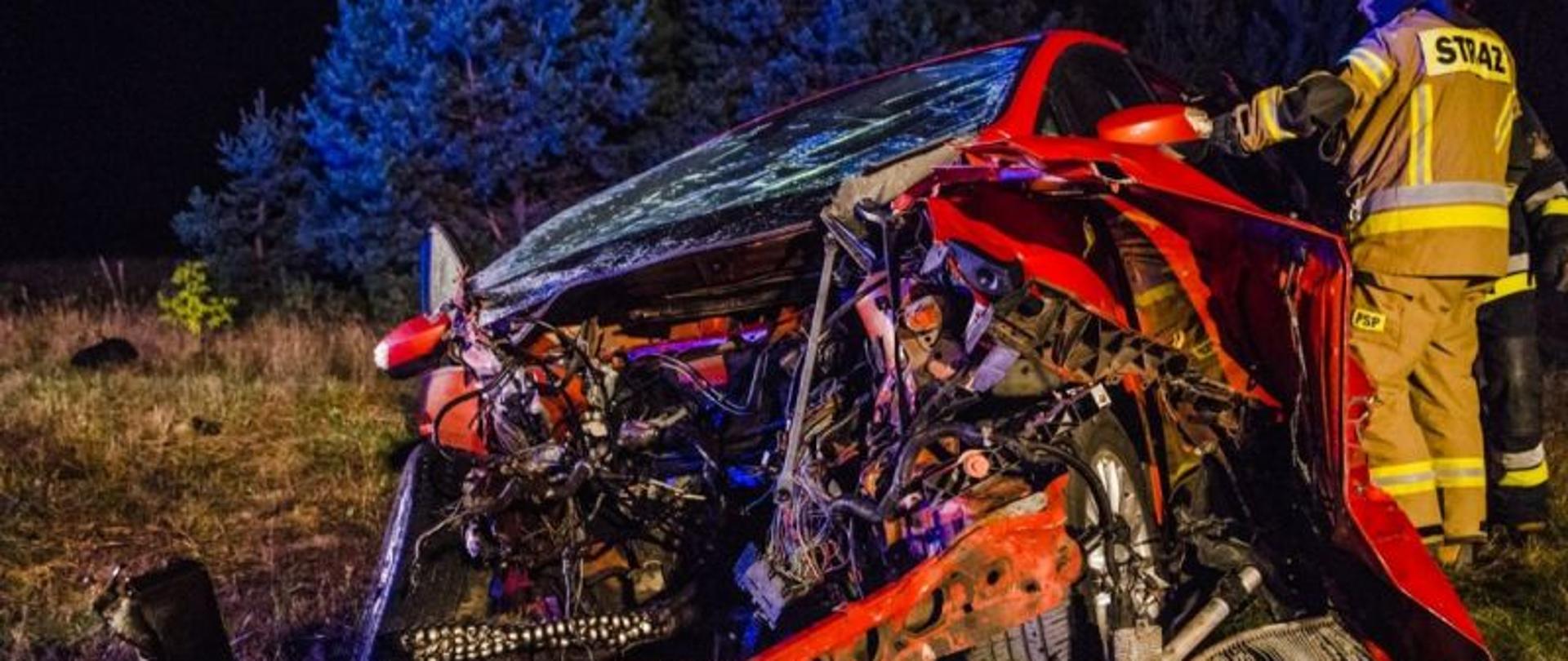 Zdjęcie przedstawia strażaka stojącego przy zniszczonym podczas wypadku aucie