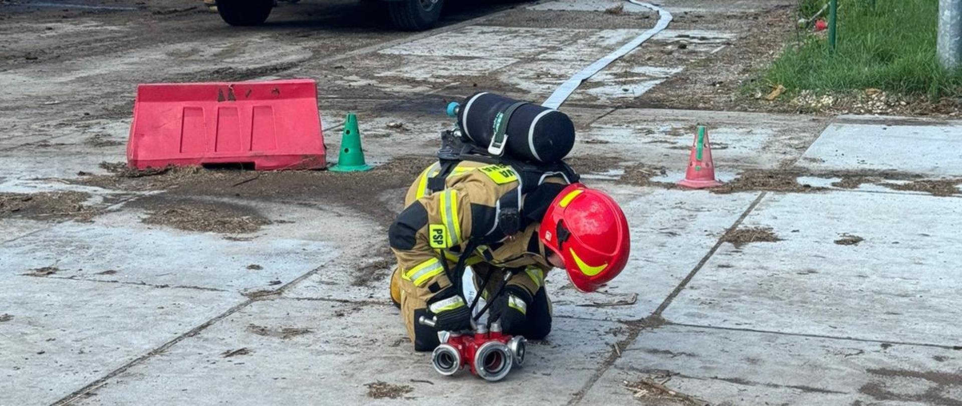 Na pierwszym planie strażak przy rozdzielaczu wodnym, w tle pozostali ratownicy i hałda odpadów