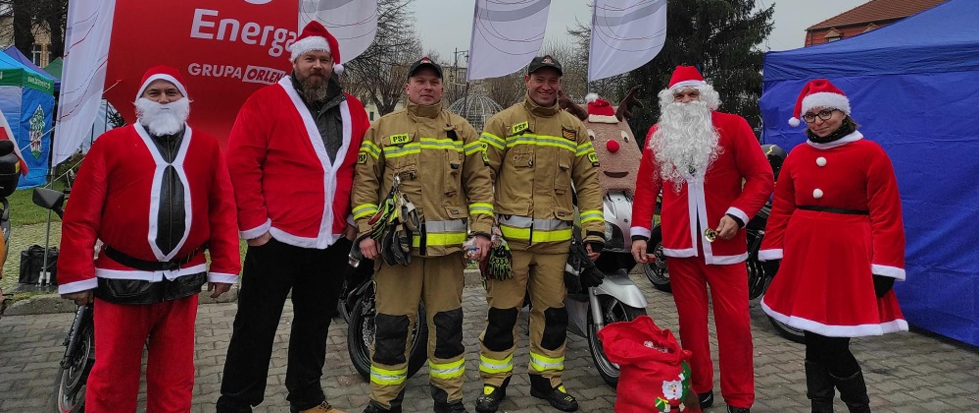  NA środku Dwóch Strażaków w mundurach, po bokach cztery osoby przebrane za mikołaje.. Z tyłu flagi.