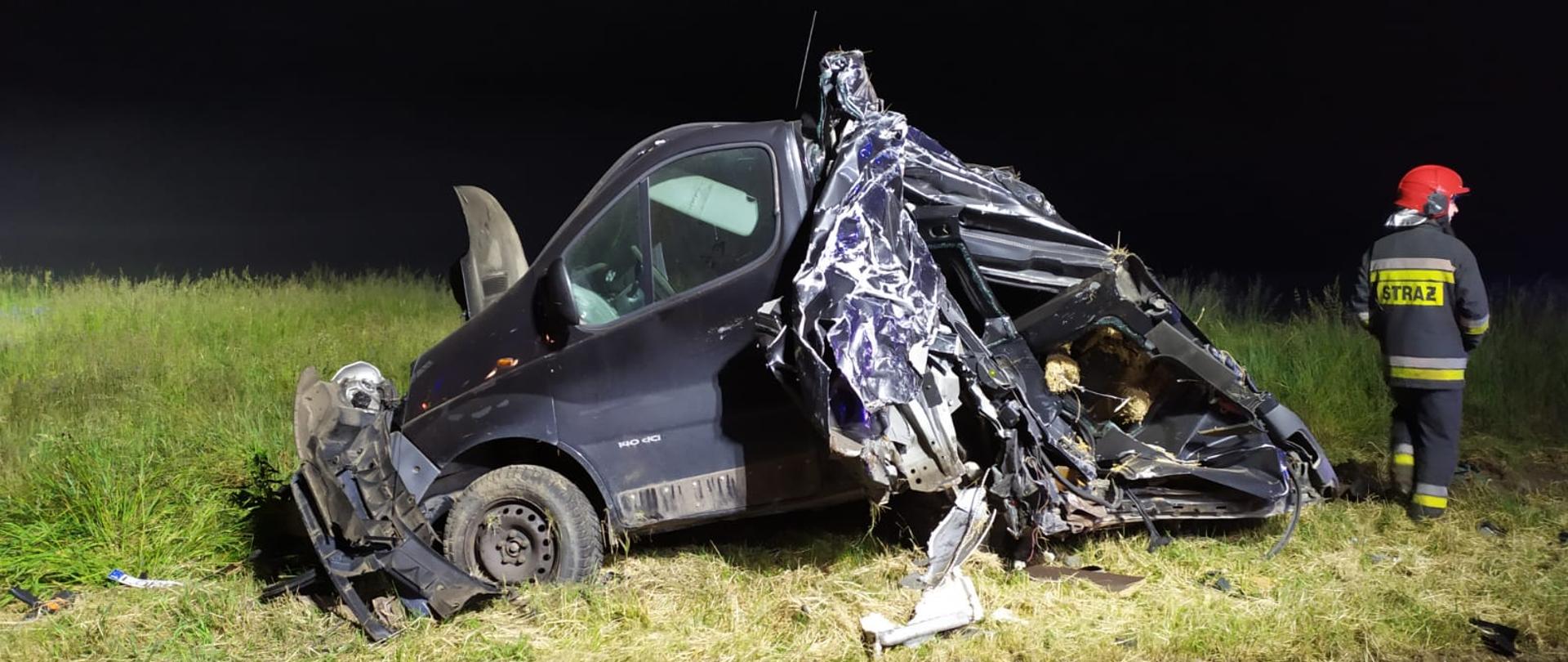 Zdjęcie przedstawia zniszczony w wyniku uderzenia samochód osobowy stojący na poboczu. Po prawej stronie widać pojedynczego strażaka.