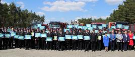 Grupowe zdjęcie druhów, parlamentarzystów i symbolicznych voucherów trzymanych w dłoniach przez kilkudziesięciu druhów. W tle pojazdy pożarnicze.