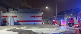 Hala zakładu produkcyjnego. Przed halą zaparkowane wozy strażackie z włączonymi sygnałami świetlnymi koloru niebieskiego. Jest ciemno. Przed zakładem świecą się lampy oświetleniowe.