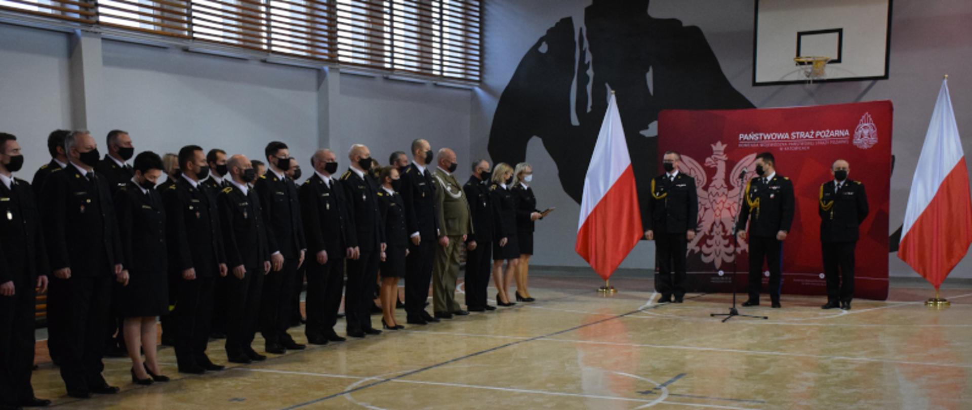 Po prawej stronie na tle napisu Państwowa Straż Pożarna Śląski Komendant Wojewódzki PSP oraz zastępcy Komendanta Wojewódzkiego, po lewej stojący w dwurzędzie funkcjonariusze ubrani w mundury galowe. 