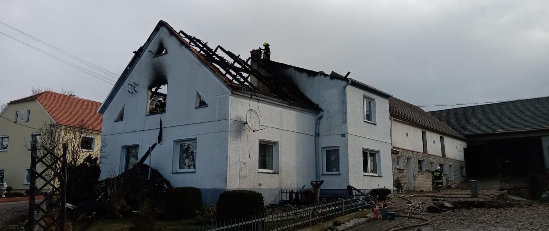 Zdjęcie przedstawia dom jednorodzinny w trakcie działań gaśniczych w miejscowości Śmicz. Wewnątrz obiektu, na strychu widoczni strażacy. Dach spalony.