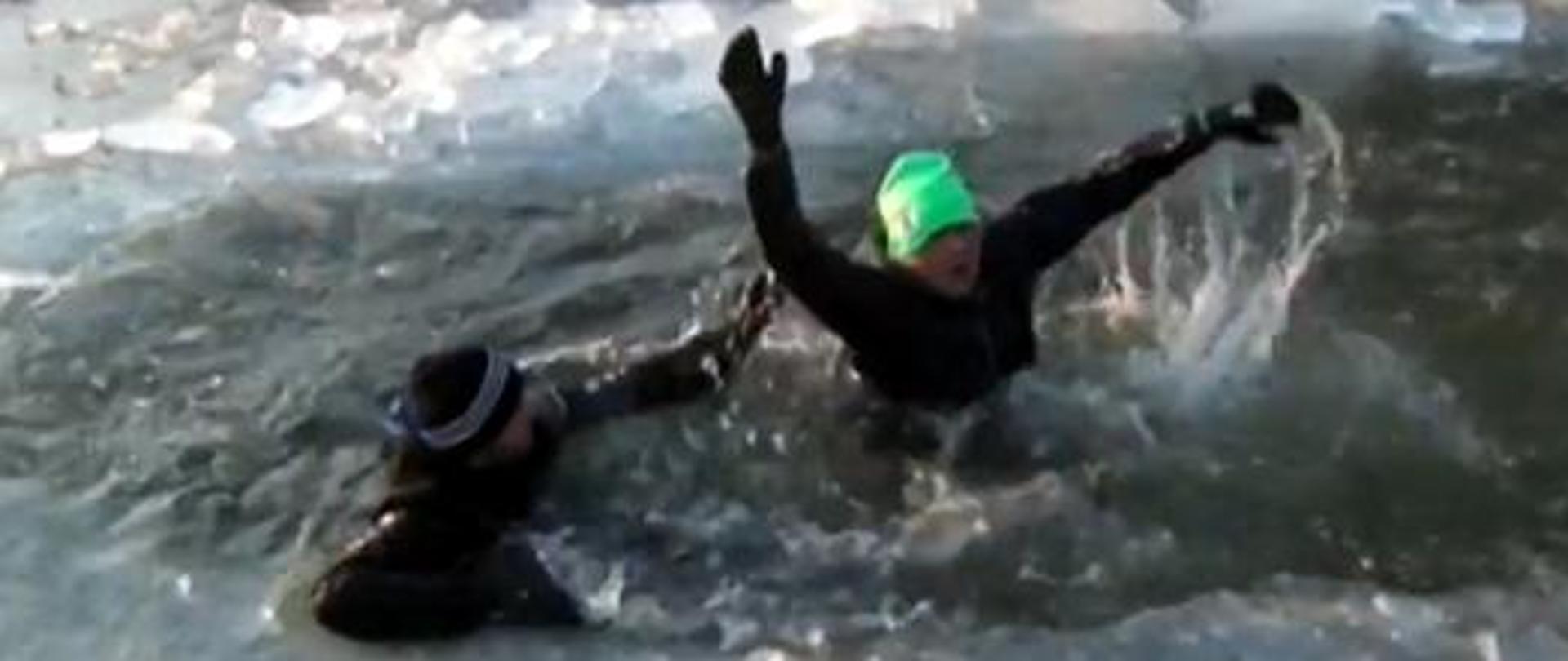 Dwóch ludzi tonie w wodzie po tym jak załamał się lód. Wymahują, rękami wzywając pomocy. Oboje ubrani na czarno. Jeden z mężczyzn ma zieloną czapkę. 