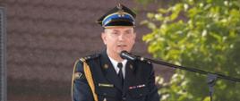 Komendant Powiatowy PSP w Ciechanów przemawia przy mównicy, trzymając dłoń na jej krawędzi. Za nim widać rozmyte tło z zielonymi drzewami.
