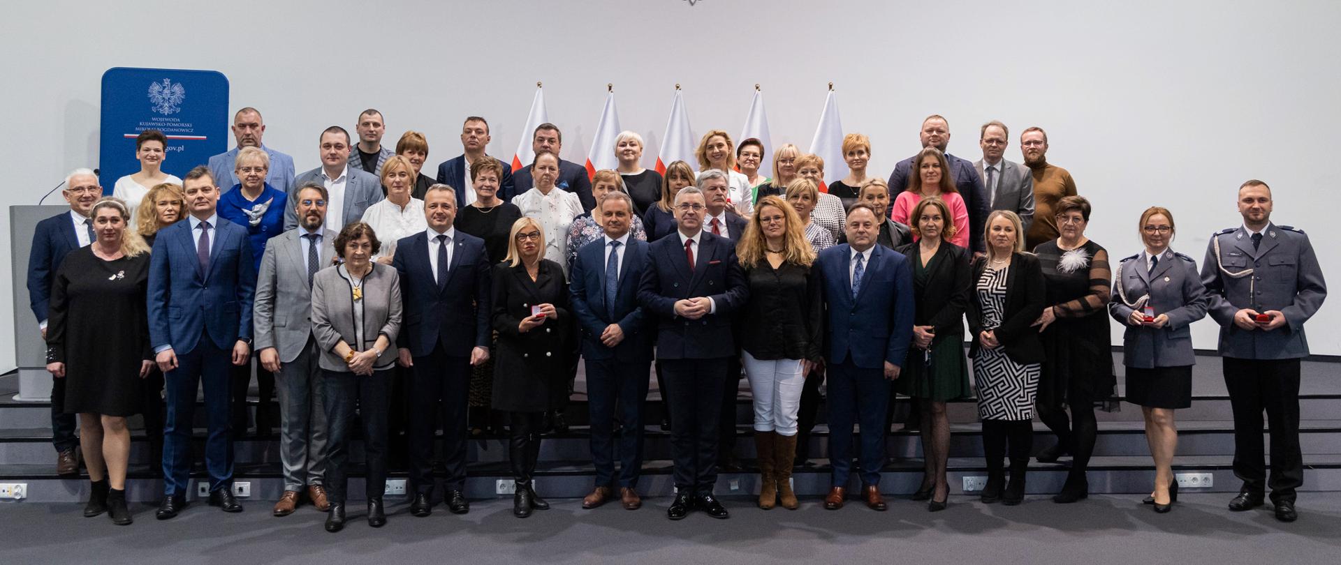 
Grupa ludzi w sali konferencyjnej stojąca na tle biało-czerwonych flag i polskiego godła
