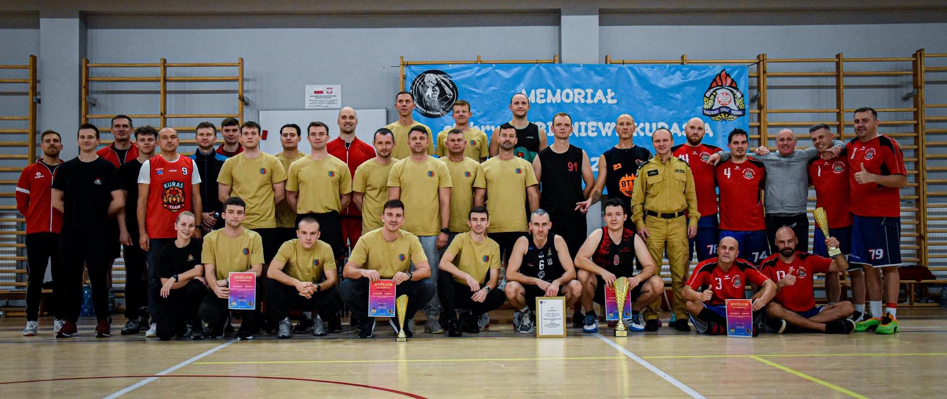 Wspólne zdjęcie uczestników Memoriału st. bryg. Zbigniewa Kurasia w Piłce Koszykowej. Na pierwszym planie zawodnicy trzymają puchary i dyplomy, a za nimi stoją drużyny w czerwonych, czarnych i beżowych koszulkach oraz przedstawiciele organizatorów.