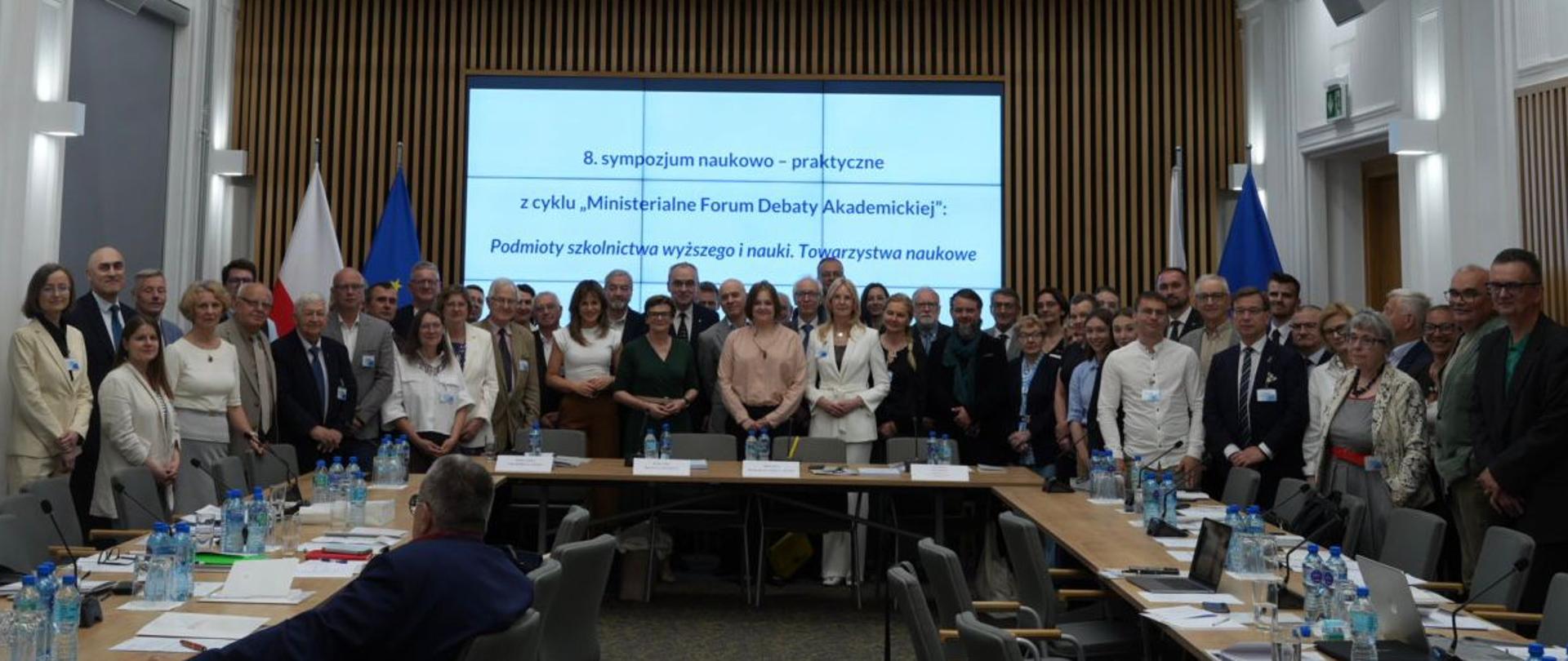 Zdjęcie zbiorowe uczestników spotkania, pod wbudowanym w ścianę z cienkich pionowych listewek wielkim ekranem z napisem 8. sympozjum naukowo-praktyczne w ramach cyklu „Ministerialne Forum Debaty Akademickiej” stoi kilkadziesiąt osób, pośrodku grupy wiceminister Zioło-Pużuk.
