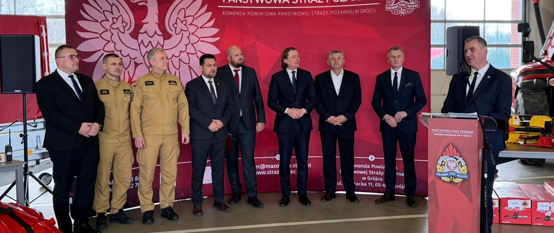 Grupa mężczyzn – część w mundurach strażackich, część w ciemnych garniturach – stoi przed czerwonym tłem z dużym napisem „Państwowa Straż Pożarna” oraz symbolem orła. Wygląda to na oficjalne wydarzenie, możliwe że konferencję, podpisanie dokumentów lub uroczyste spotkanie. Jeden z nich po prawej stronie stoi przy mównicy i przemawia.
Całość przedstawia formalne spotkanie służb pożarniczych i lokalnych przedstawicieli władz w Grójcu.
