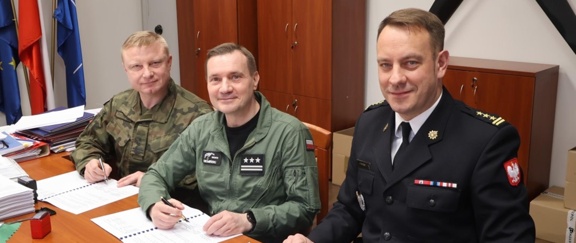Zdjęcie wykonane w jednym z pomieszczeń bazy wojskowej. Na zdjęciu widać biurko. Na biurku leżą trzy dokumenty zawierające treść porozumienia o współpracy. Przy biurku od lewej strony siedzą: Szefem Delegatury Wojskowej Ochrony Przeciwpożarowej w Warszawie, Dowódca 42 Bazy Lotnictwa Szkolnego w Radomiu oraz Komendant Miejski Państwowej Straży Pożarnej w Radomiu. Mężczyźnie ubrani są w umundurowanie właściwe do reprezentowanych przez siebie formacji. W rękach trzymają długopisy, którymi złoża swoje podpisy pod dokumentem. 