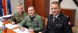 Zdjęcie wykonane w jednym z pomieszczeń bazy wojskowej. Na zdjęciu widać biurko. Na biurku leżą trzy dokumenty zawierające treść porozumienia o współpracy. Przy biurku od lewej strony siedzą: Szefem Delegatury Wojskowej Ochrony Przeciwpożarowej w Warszawie, Dowódca 42 Bazy Lotnictwa Szkolnego w Radomiu oraz Komendant Miejski Państwowej Straży Pożarnej w Radomiu. Mężczyźnie ubrani są w umundurowanie właściwe do reprezentowanych przez siebie formacji. W rękach trzymają długopisy, którymi złoża swoje podpisy pod dokumentem. 