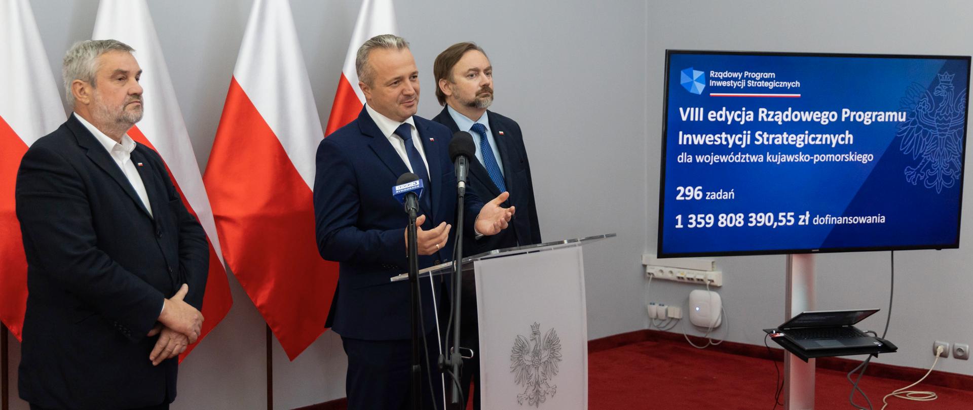 
3 mężczyzn w garniturach stających przy mikrofonie w sali konferencyjnej
