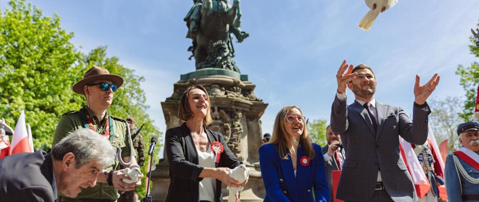 Kilka osób stoi pod pomnikiem Jana II Sobieskiego i wypuszcza z rąk białe gołębie