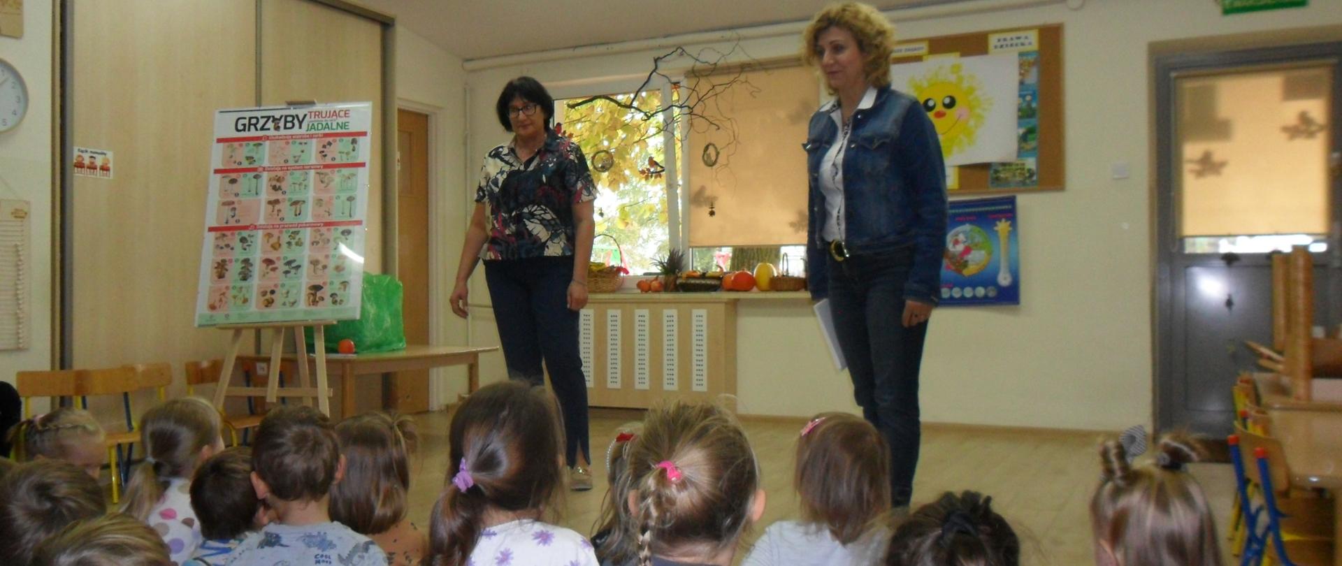 Przedstawiciele Państwowego Powiatowego Inspektora Sanitarnego w Garwolinie w czasie pogadanki z przedszkolakami na sali. W tle plakat "Grzyby trujące i jadalne".