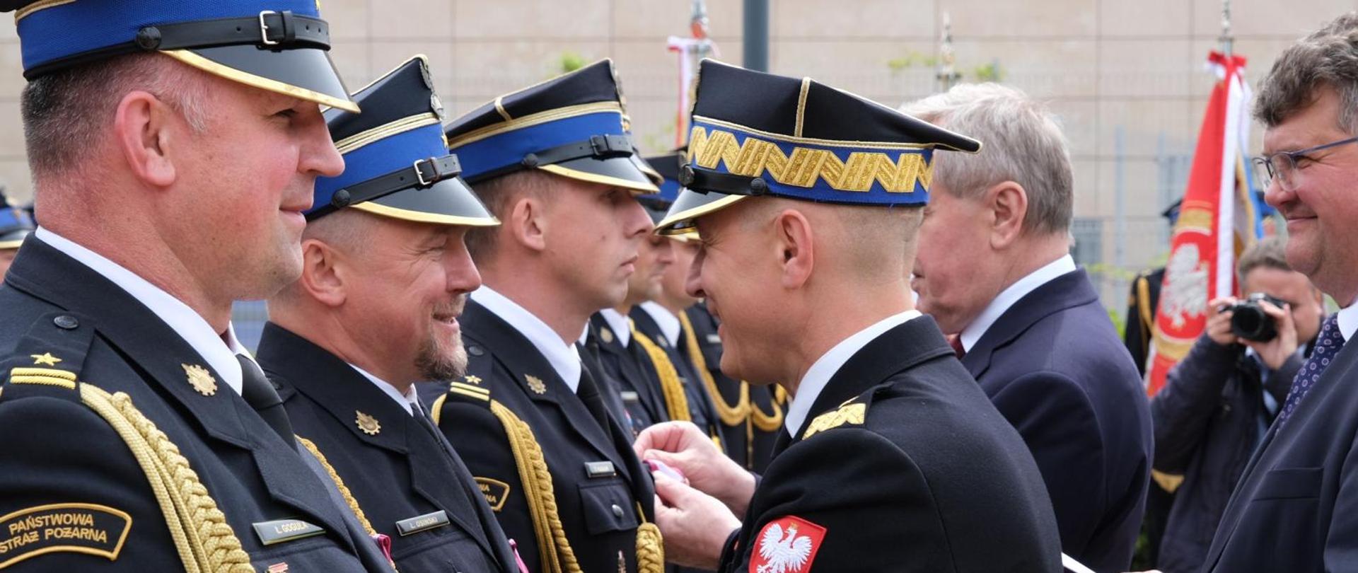Komendant główny PSP w trakcie wręczania odznaczeń oficerom PSP stojącemu w szeregu. W tle pozostali uczestnicy uroczystości i poczet sztandarowy. 