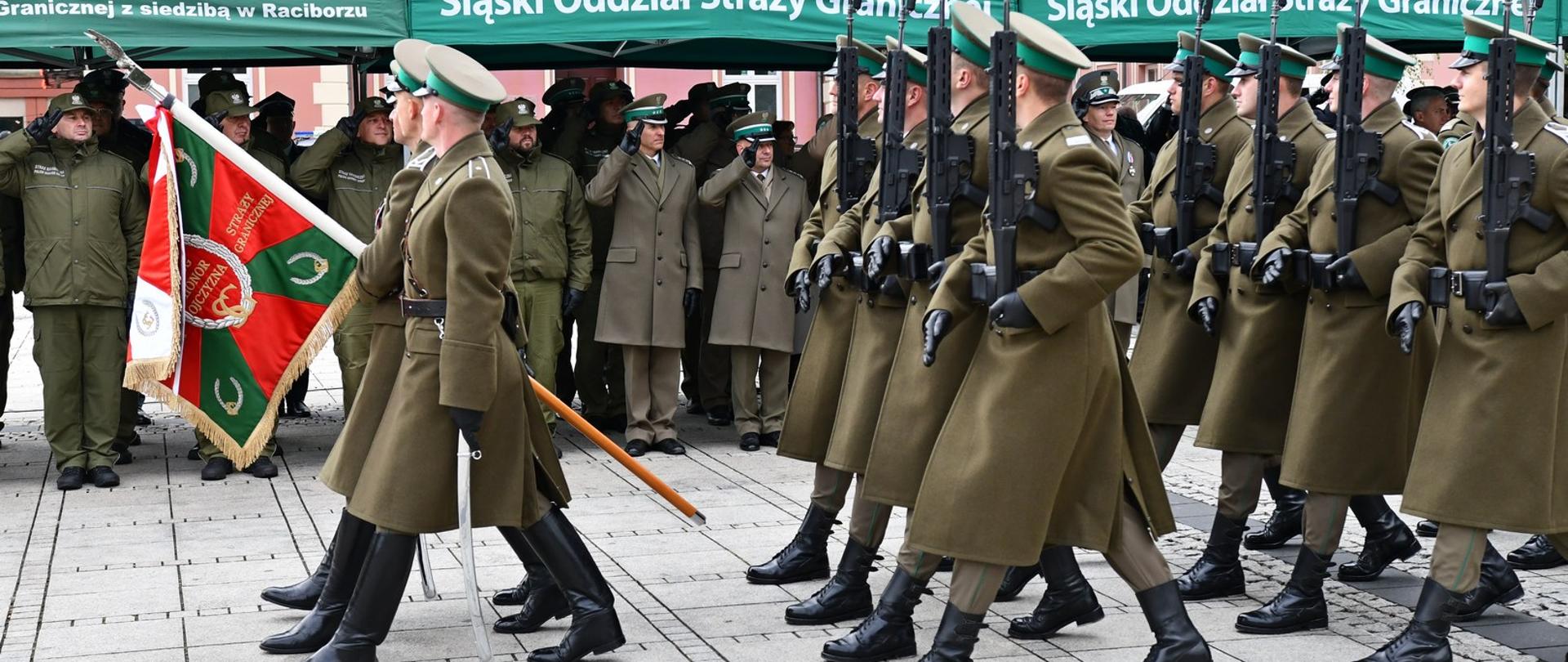 Maszerująca kompania honorowa Straży Granicznej na czele z pocztem sztandarowym, w tle salutujący funkcjonariusze Straży Granicznej obecni na uroczystych obchodach rocznicowych
