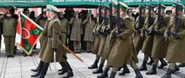 Maszerująca kompania honorowa Straży Granicznej na czele z pocztem sztandarowym, w tle salutujący funkcjonariusze Straży Granicznej obecni na uroczystych obchodach rocznicowych