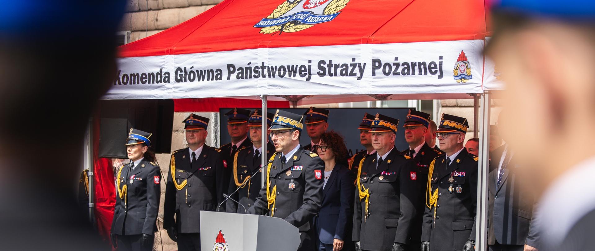 Komendant Główny Państwowej Straży Pożarnej przemawia przy mównicy pod namiotem Komendy Głównej PSP. Za nim stoją funkcjonariusze PSP w mundurach galowych oraz zaproszeni goście.