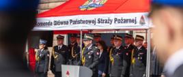 Komendant Główny Państwowej Straży Pożarnej przemawia przy mównicy pod namiotem Komendy Głównej PSP. Za nim stoją funkcjonariusze PSP w mundurach galowych oraz zaproszeni goście.