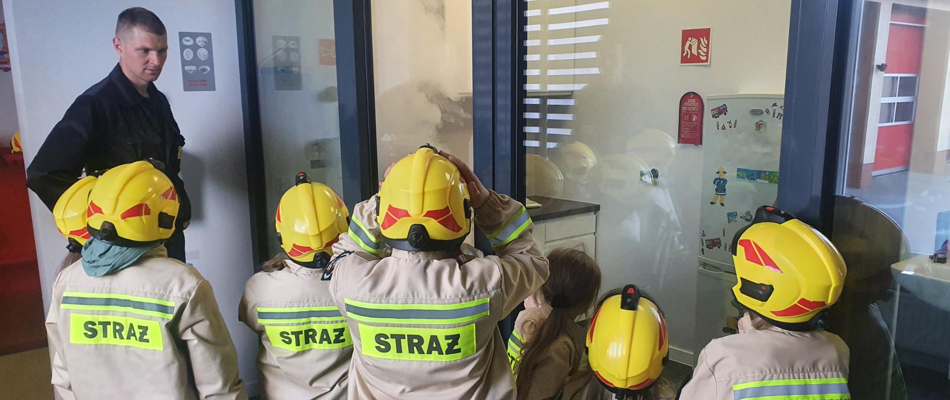 Grupa dzieci w kurtkach strażackich i hełmach w trakcie zajęć na Sali Edukacyjnej.