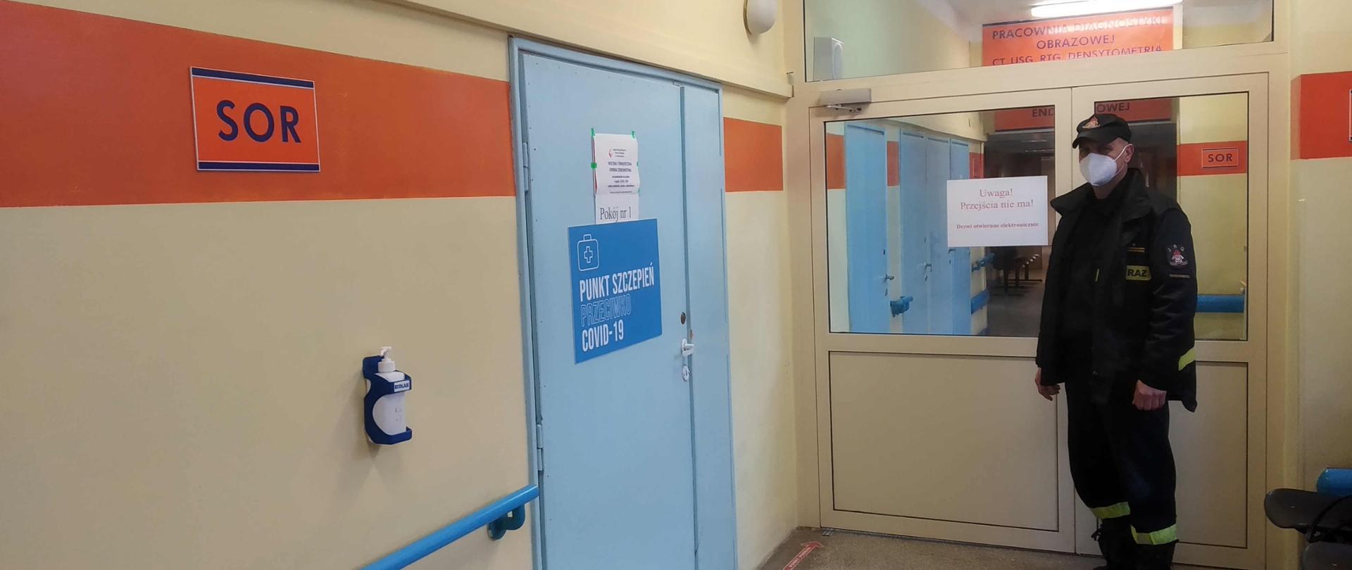 Zdjęcie zrobione na szpitalnym korytarzu. Po lewej stronie znajdują się niebieski drzwi z napisem punkt szczepień przeciwko covid-19. Naprzeciwko drzwi stoi strażak ubrany w ubranie koszarowe, który oczekuje na szczepienie. Twarz ma zasłoniętą maseczką. Za nim znajdują się oszklone drzwi prowadzące do dalszej części szpitala.