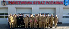 Strażacy stojący do zdjęcia grupowego na tle Komendy PSP w Pleszewie