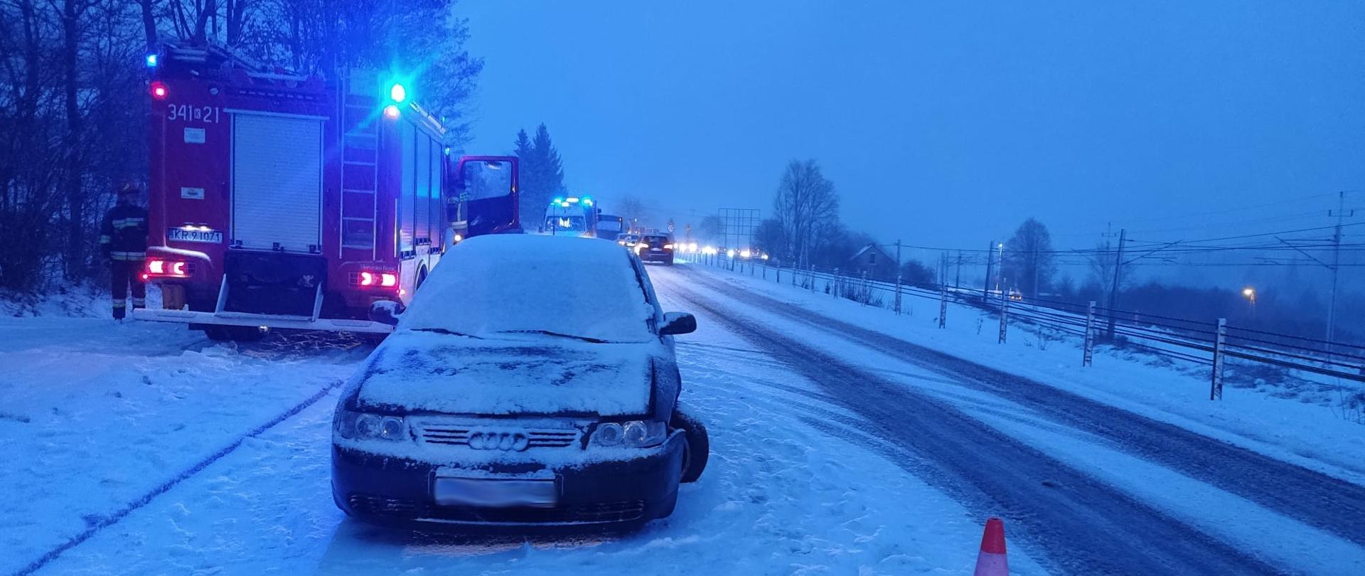 Uszkodzony samochód osobowy na poboczu a obok niego samochód pożarniczy. Droga pokryta śniegiem.