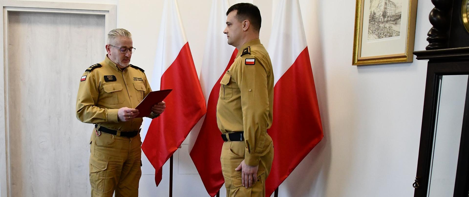 Strażak w okularach w mundurze koloru musztardowego w koszuli trzyma teczkę przed nim stoi funkcjonariusz Państwowej Straży Pożarnej w mundurze koloru musztardowego za nimi ustawione są trzy flagi Polski na ścianie wisi obraz oraz przed nimi stoi zegar.