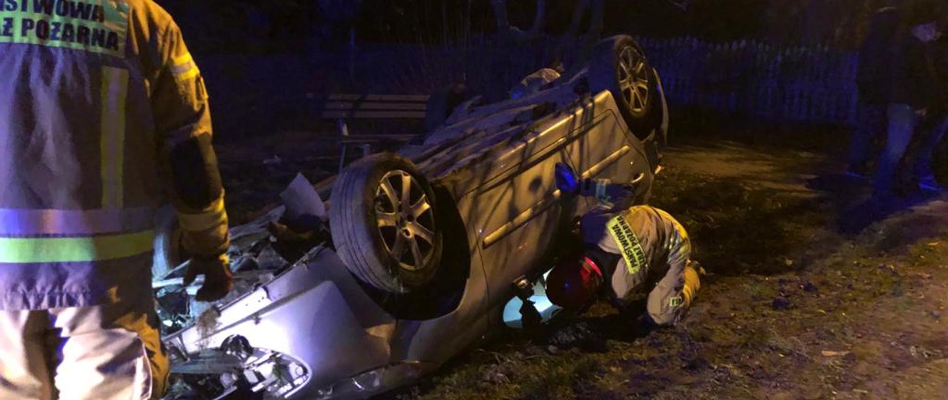 Samochód osobowy leży na dachu. Jeden ze strażaków zagląda do środka czy ktoś jest w środku. Drugi stoi z boku. 