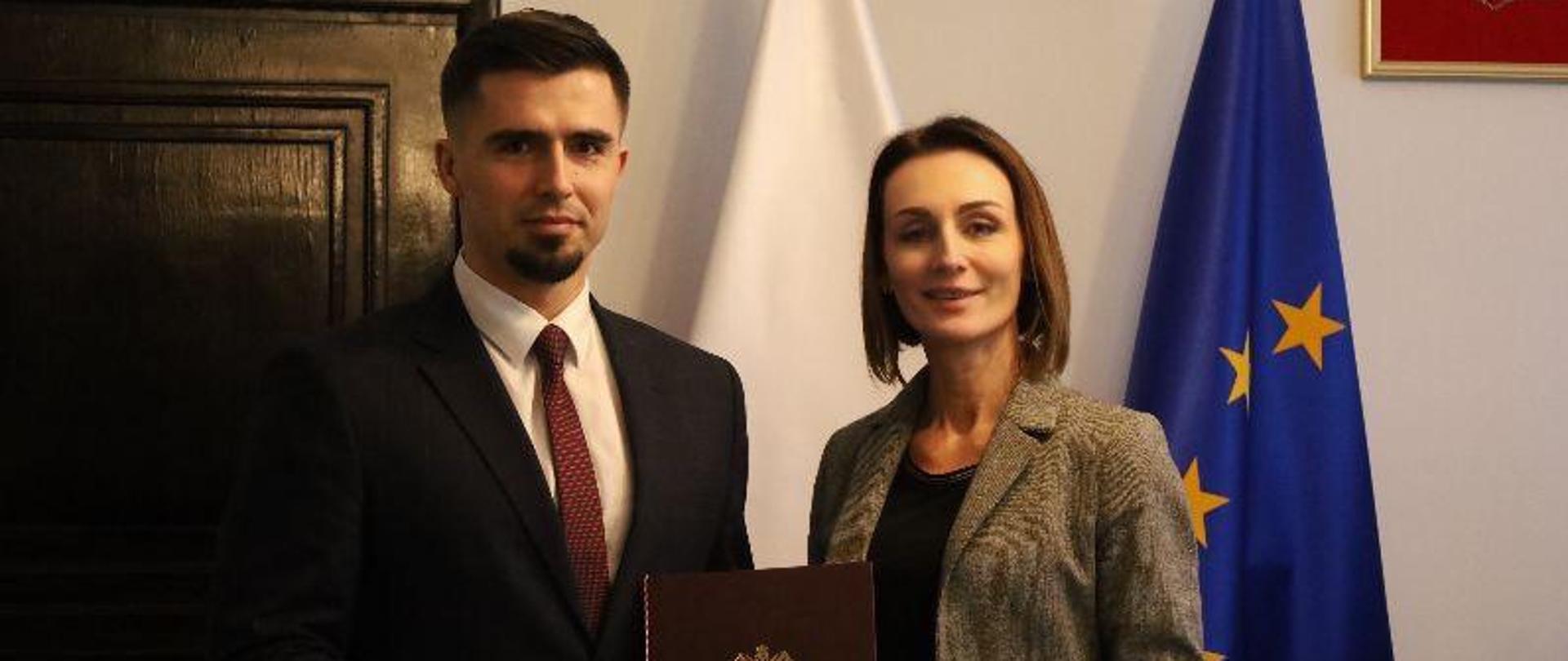 Kobieta ( wojewoda) stojąca na tle fagi państwowej i unijnej wręcza powołanie na pełniącego obowiązki wójta gminy Pruszcz Gdański mężczyźnie. Gabinet wojewody pomorskiej