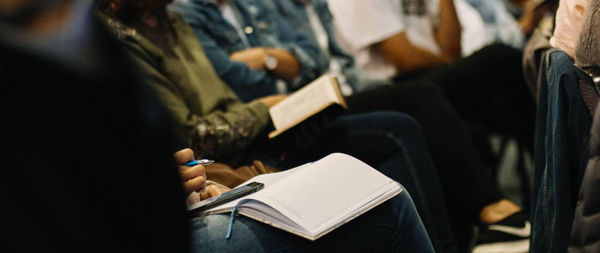 fotografia przedstawia sylwetki osób siedzących w rzędzie, które trzymają notatniki na kolanach