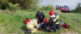 Zdjęcie przedstawia scenę udzielania przez 4 strażaków pierwszej pomocy przedmedycznej osobie wyciągniętej z rzeki Wisły. Na dalszym planie widoczny czerwony samochód pożarniczy oraz łódź ze strażakami.