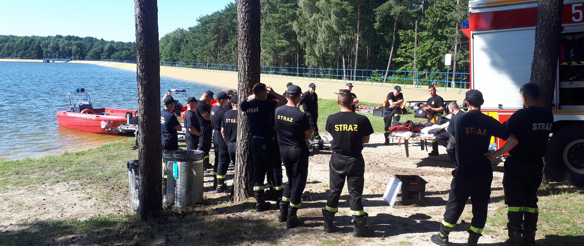 Zdjęcie przedstawia ćwiczenia JRG Ostrzeszów na zbiorniku wodnym Blewązka z Grupą Specjalistyczną Ratownictwa Wodno - Nurkowego z Sieradza. Na zdjęciu widzimy funkcjonariuszy, którzy przygotowują się do ćwiczeń w wodzie. Zdjęcie wykonane w porze dziennej na terenie zalewu.