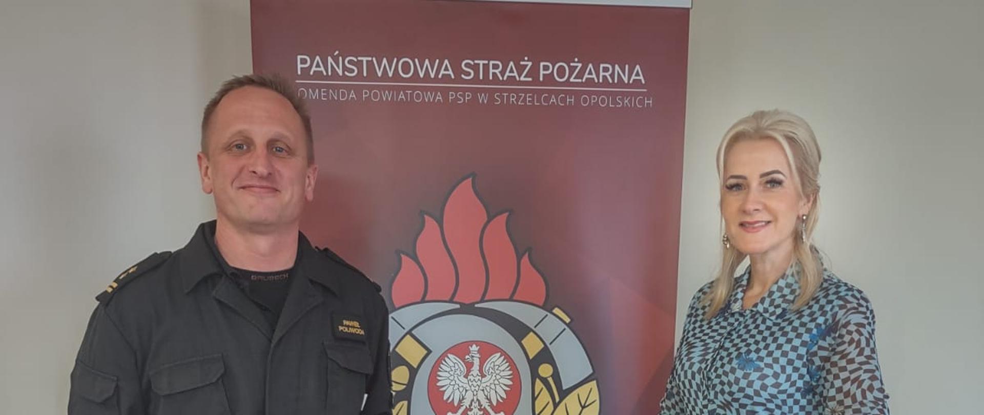 Na zdjęciu znajduje się Strażak ubrany w umundurowanie dowódczo-sztabowe oraz kobieta, w tle stoi baner z logo PSP