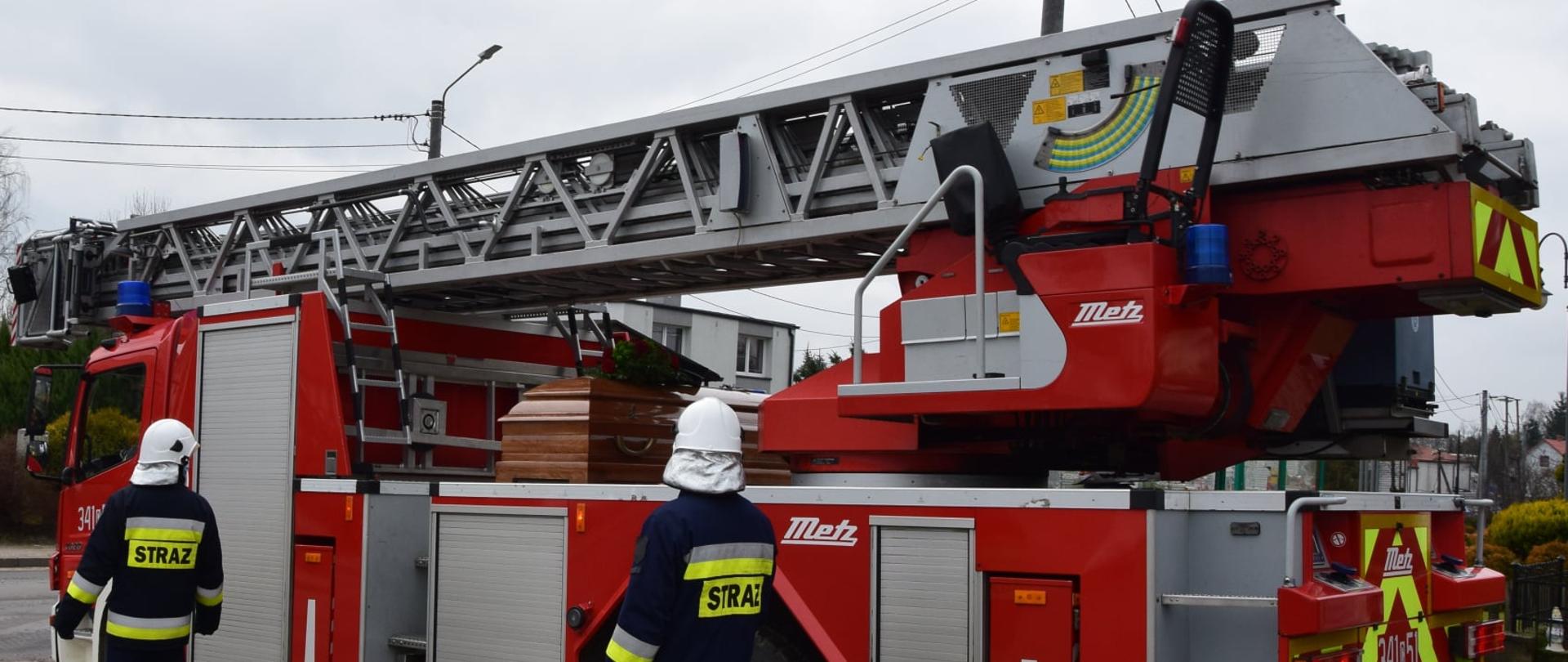 Na samochodzie strażackim z drabiną mechaniczną leży trumna w kolorze brązowym. Przy samochodzie w asyście idzie dwóch strażaków w ubraniach specjalnych w hełmach na głowie.