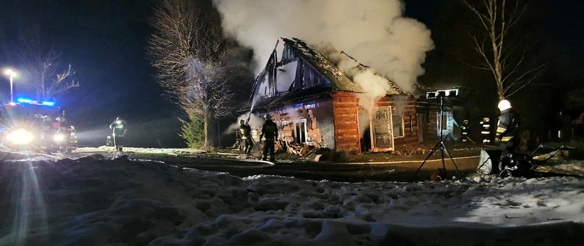 Kolorowa fotografia wykonana w porze nocnej. Przedstawia gaszenie pożaru drewnianego budynku niezamieszkałego. Na pierwszym planie widoczny leżący śnieg. Na drugim planie częściowo zniszczony przez ogień budynek mieszkalny, z którego wydobywa się dużo dymu. Wokół liczni strażacy gaszący pożar budynku. Po lewej widoczny pojazd gaśniczy straży pożarnej oświetlający miejsce pożaru. W tle ciemne niebo.