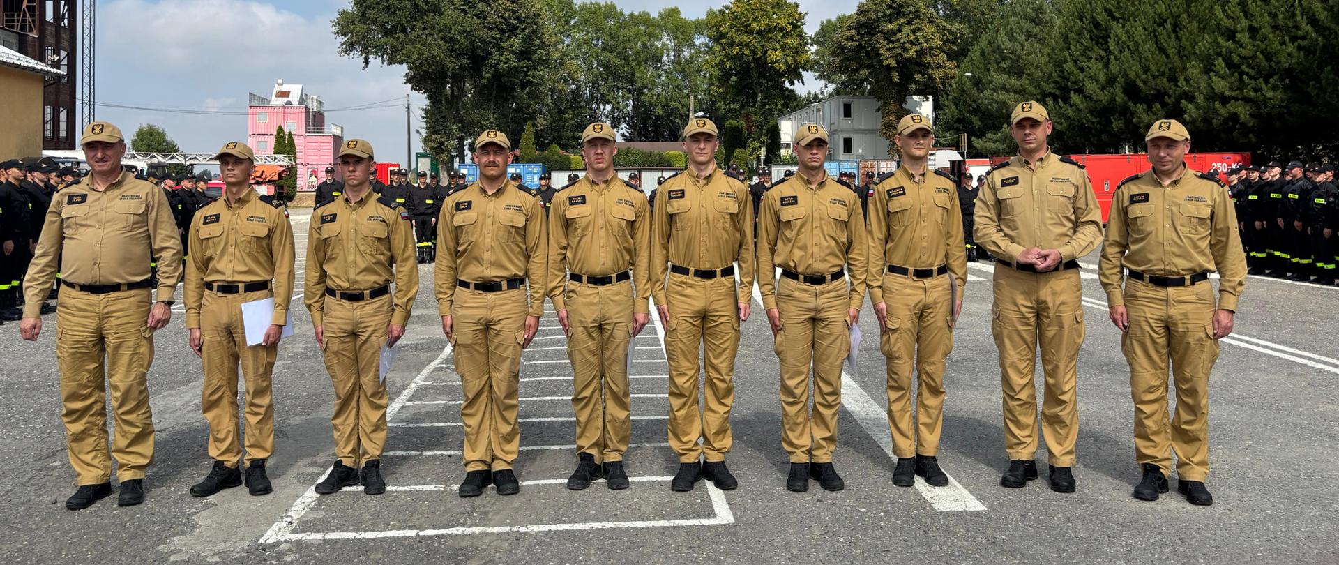 5. Na zdjęciu widoczny jest rząd funkcjonariuszy w piaskowych mundurach służbowych. Siedmiu z nich trzyma białe teczki. Wszyscy stoją w postawie zasadniczej. W tle znajdują się funkcjonariusze w czarnych mundurach służbowych, ustawieni w równych dwuszeregach, a także widoczna jest infrastruktura poligonu.
